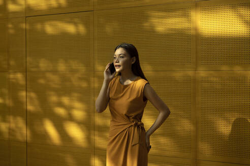 Beautiful woman talking through smart phone while standing with hand on hip in front of yellow wall - EAF00037