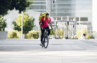 Lächelnder Zusteller mit gelbem Rucksack auf dem Fahrrad in der Stadt - JCCMF03486