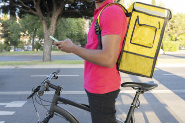 Zusteller mit gelbem Rucksack, der sein Smartphone benutzt, während er mit dem Fahrrad steht - JCCMF03498