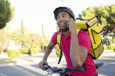 Lächelnder Zusteller, der sich auf ein Fahrrad stützt und mit seinem Smartphone spricht - JCCMF03500