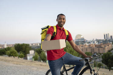 Lächelnder Zusteller, der ein Paket übergibt, während er auf dem Fahrrad sitzt - JCCMF03523