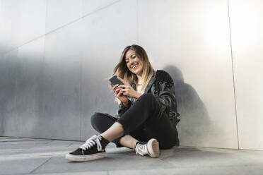 Smiling woman using smart phone in front of silver wall - MTBF01068