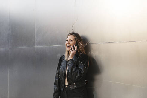 Junge Frau, die vor einer silbernen Wand mit einem Smartphone spricht - MTBF01073