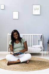 Lächelnde schwangere junge Frau im Schneidersitz mit Buch im Schlafzimmer - GIOF13152