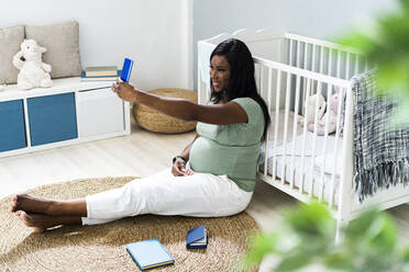 Lächelnde schwangere junge Frau, die ein Selfie macht, während sie vor der Krippe zu Hause sitzt - GIOF13160