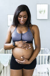 Lächelnde schwangere Frau, die im Schlafzimmer ihren Unterleib berührt - GIOF13162