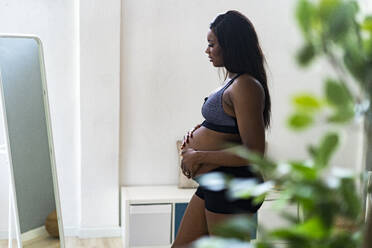 Schwangere Frau, die ihren Unterleib berührt, während sie im Schlafzimmer vor einem Spiegel steht - GIOF13164