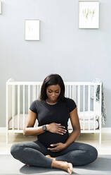 Schwangere Frau, die ihren Bauch berührt, während sie im Schneidersitz auf einer Übungsmatte im Schlafzimmer sitzt - GIOF13167