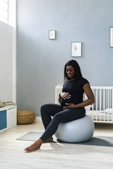 Smiling pregnant woman touching abdomen while sitting on fitness ball at home - GIOF13177