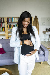 Glückliche junge schwangere Freiberuflerin, die ihren Unterleib berührt, während sie zu Hause steht - GIOF13198