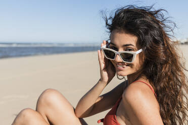 Smiling young woman wearing sunglasses while sitting at beach - JRVF01394