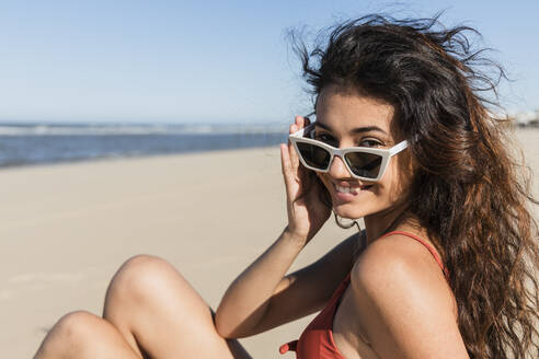 Lächelnde junge Frau mit Sonnenbrille, die am Strand sitzt - JRVF01394