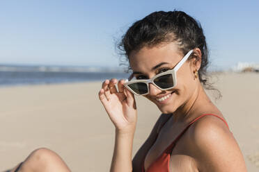 Young woman wearing sunglasses sitting at beach on sunny day - JRVF01395