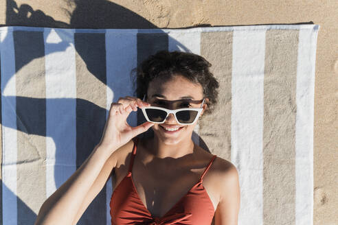 Lächelnde junge Frau mit Sonnenbrille, die auf einem Handtuch am Strand liegt - JRVF01397