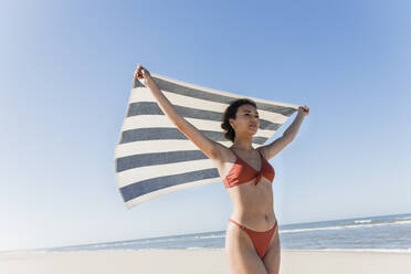 Junge Frau winkt mit Handtuch am Strand im Urlaub - JRVF01399