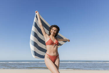 Schöne junge Frau mit erhobenen Armen hält Handtuch am Strand - JRVF01401