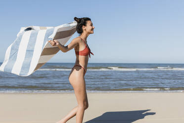 Young woman waving towel while running at beach during vacation - JRVF01402