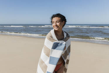 Smiling woman wrapped in towel looking away while standing at beach - JRVF01405