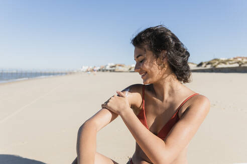 Smiling young woman applying sunscreen while sitting at beach on sunny day - JRVF01409