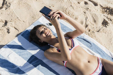Lächelnde junge Frau, die ein Mobiltelefon benutzt, während sie am Strand entspannt - JRVF01411