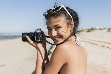 Smiling young woman with camera standing at beach on sunny day - JRVF01418