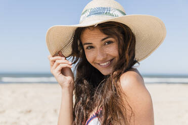 Lächelnde Frau mit Sonnenhut am Strand im Urlaub - JRVF01420