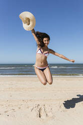 Carefree woman with hat jumping at beach during vacation - JRVF01423