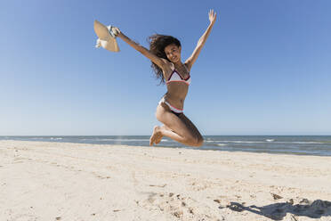 Carefree woman with arms raised jumping at beach - JRVF01424