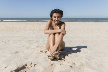 Smiling young woman hugging knees on sand at beach - JRVF01426