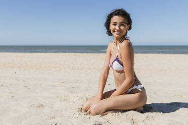 Smiling beautiful woman kneeling on sand at beach during sunny day - JRVF01428