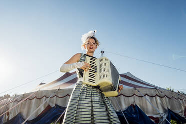 Weibliche Darstellerin spielt Akkordeon vor einem Zirkuszelt - MEUF03839
