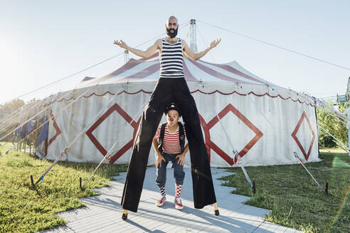 Männlicher Clown stehend mit Artisten auf Stelzen vor einem Zirkuszelt - MEUF03846