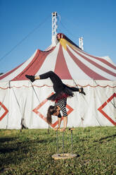 Weiblicher Akrobat im Handstand auf einem Stock vor einem Zirkuszelt - MEUF03865