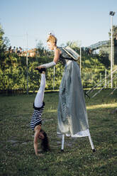 Female artist helping acrobat doing handstand on meadow - MEUF03877