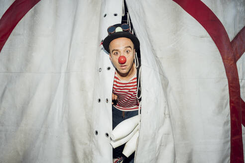 Male clown with hat and juggling pin peeking through circus tent entrance - MEUF03899