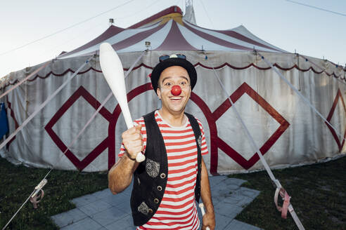 Lächelnder männlicher Clown, der einen Jonglierstift hält, während er vor einem Zirkuszelt steht - MEUF03901