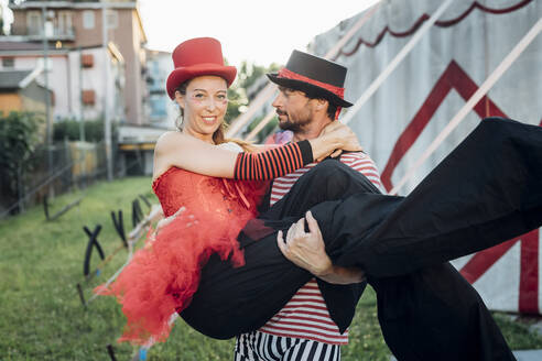 Männlicher Akrobat, der eine Artistin trägt, während er vor einem Zirkuszelt steht - MEUF03913