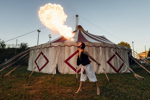 Männlicher Akrobat, der Feuer bläst, während er vor einem Zirkuszelt steht - MEUF03933