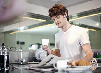 Young man reading newspaper while having breakfast in kitchen at home - AJOF01547