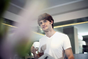 Junger Mann mit Kaffeetasse schaut in der Küche weg - AJOF01548