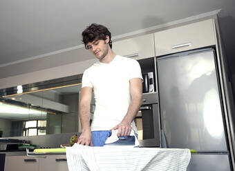 Young man ironing shirt in kitchen at home - AJOF01549