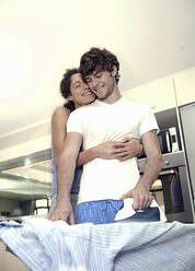 Glückliche Frau umarmt Mann, der in der Küche sein Hemd bügelt - AJOF01550