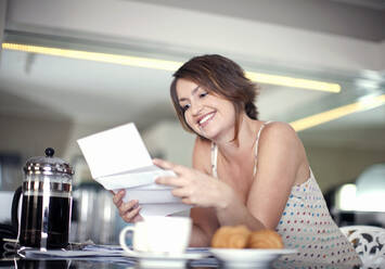 Smiling young woman reading letter while sitting at kitchen island - AJOF01555