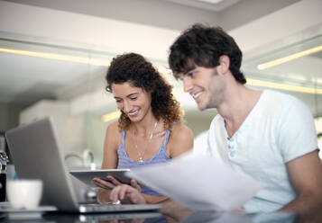 Lächelnder Mann, der einen Laptop benutzt, während seine Freundin ein Dokument in der Küche hält - AJOF01563