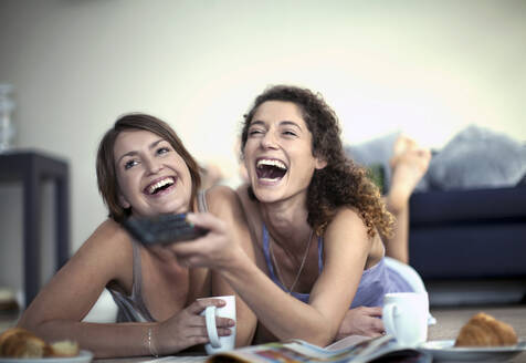 Fröhliche Frau, die mit der Fernbedienung den Sender wechselt, während sie mit einer Freundin zu Hause fernsieht - AJOF01567