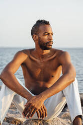Nachdenklicher Mann ohne Hemd schaut weg, während er am Strand sitzt - VPIF04558