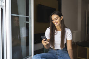 Glückliche schöne Frau, die mit ihrem Handy im Internet surft, während sie am Fenster sitzt - MTBF01076