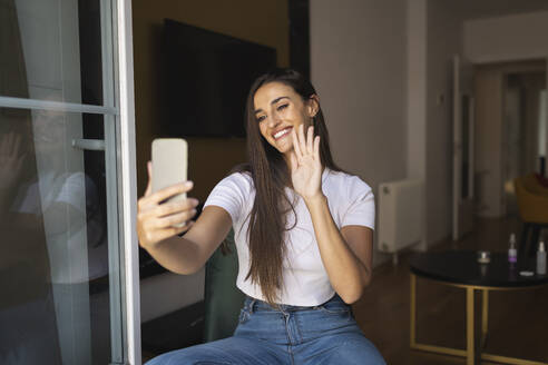 Lächelnde junge Frau winkt mit der Hand während eines Videoanrufs über ein Smartphone - MTBF01077