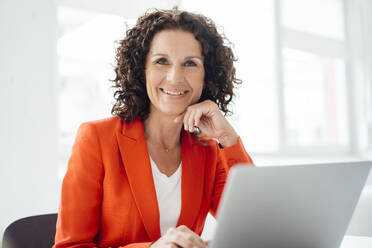 Smiling businesswoman with laptop sitting at office - MOEF03841
