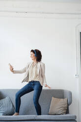 Weibliche Fachkraft hält Handy beim Tanzen auf dem Sofa im Büro - MOEF03863